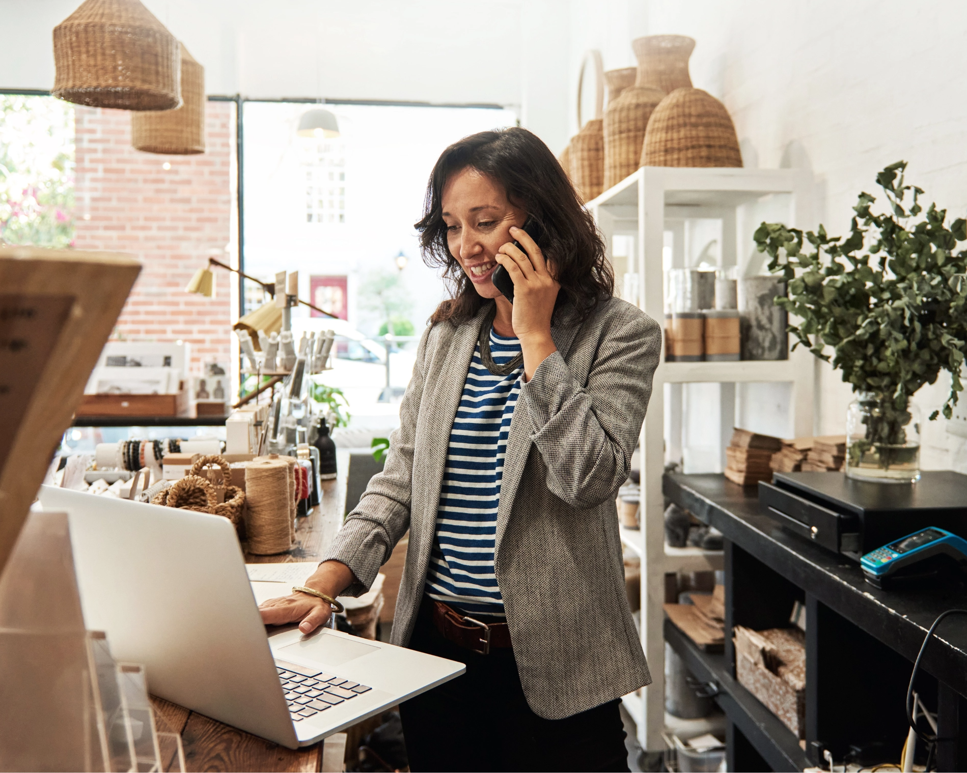 Antreprenoare zâmbind vorbind la telefon și lucrând pe laptop în magazinul său, ilustrând procesul simplu de obținere a liniei de credit pentru plăți furnizori în 3 pași: creare cont, decizie în 1 zi lucrătoare și plata directă a furnizorilor
