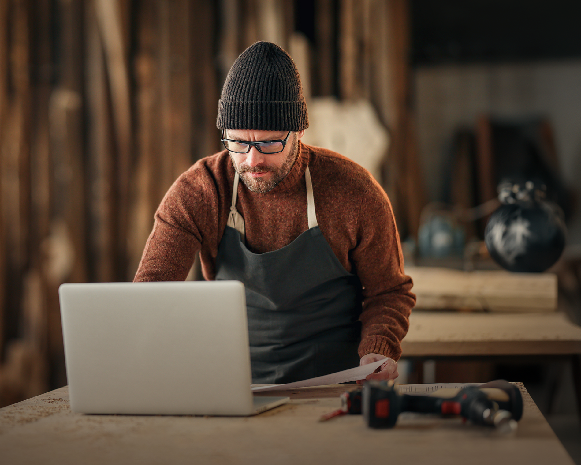 Meșter tâmplar cu șorț și căciulă lucrând pe laptop în atelier, ilustrând procesul simplu de obținere a creditului pentru afaceri în 3 pași prin Micro Credit IMM